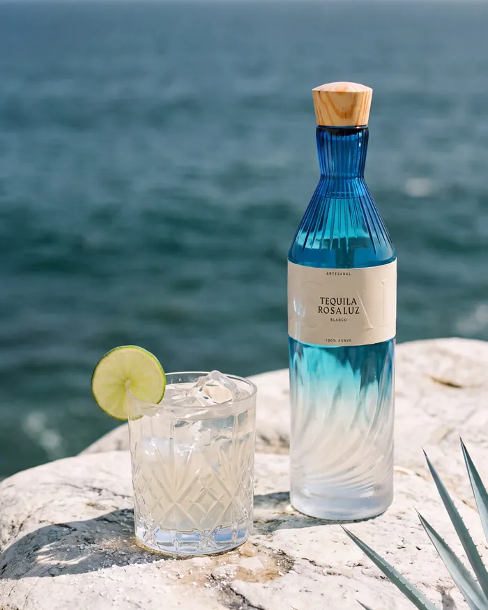 A Skinny Margarita with a lime wheel beside a bottle of Tequila Rosaluz Blanco on a rock above the sea