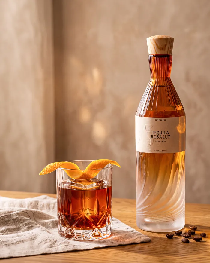 A Stevia Old Fashioned with an orange twist beside a bottle of Tequila Rosaluz Reposado on a wooden table