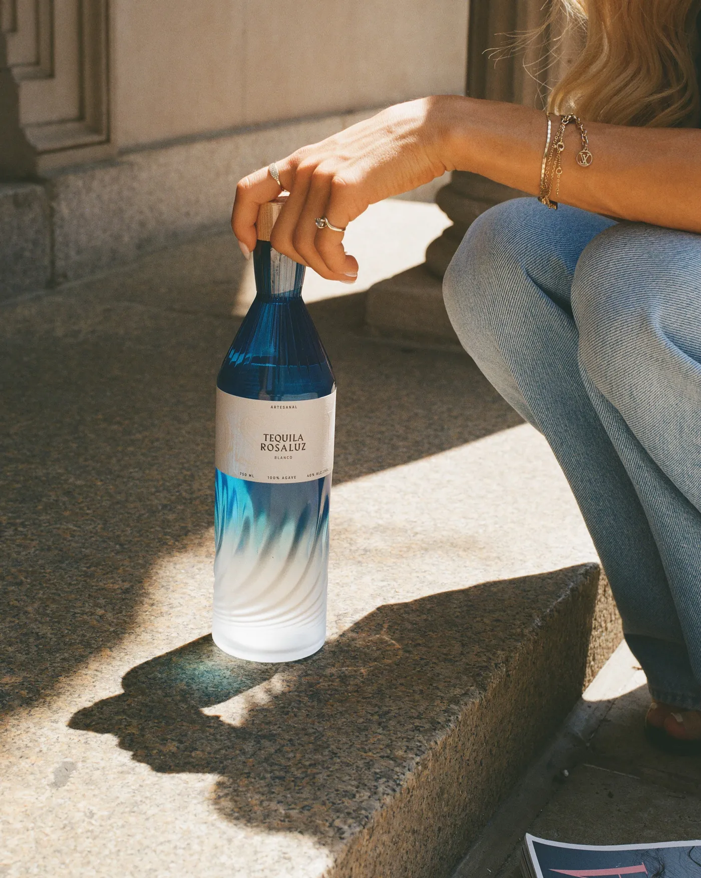 A hand resting a bottle of Tequila Rosaluz Blanco on stone steps in low sun