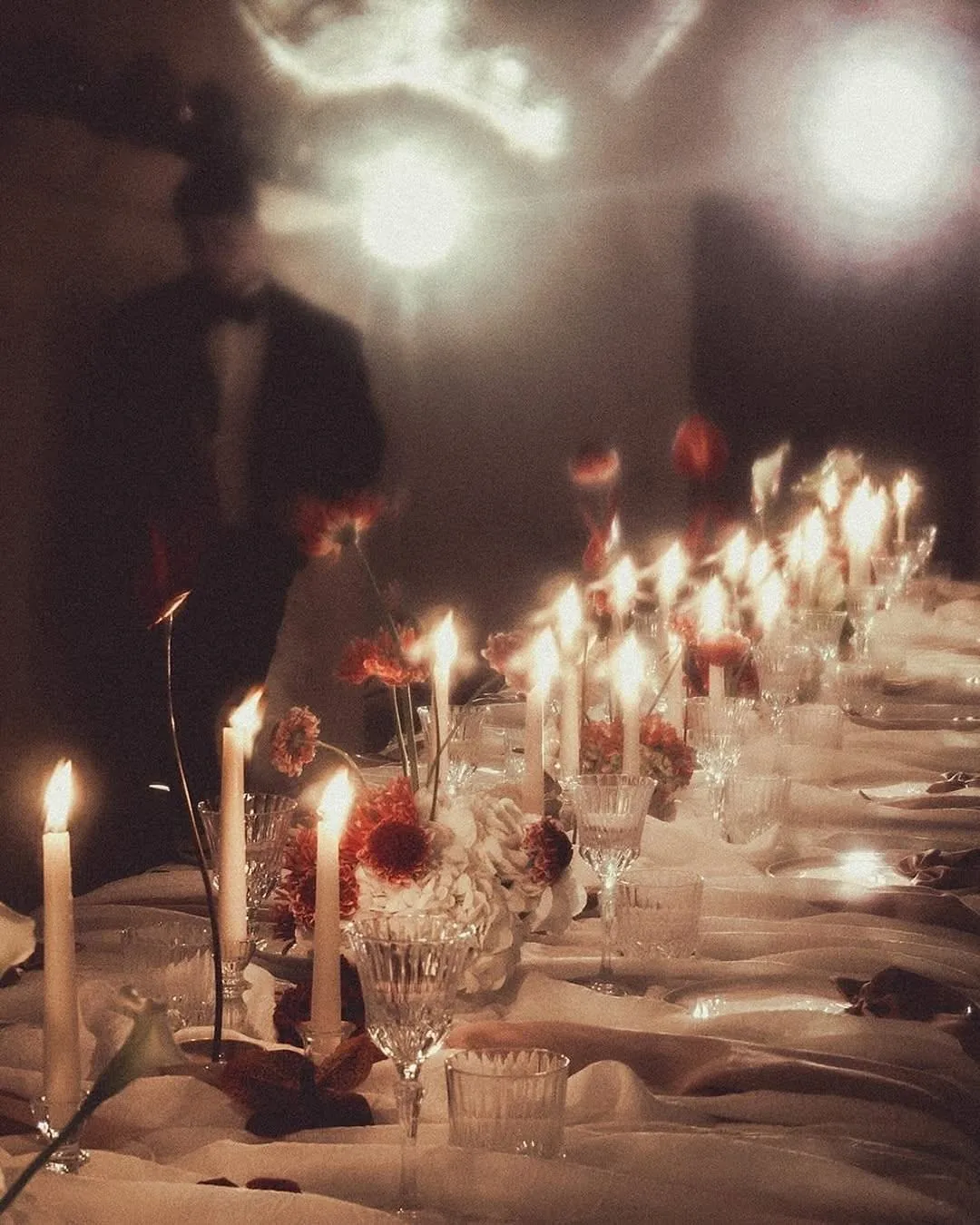 A long table set with candles and flowers, guests arriving in low light